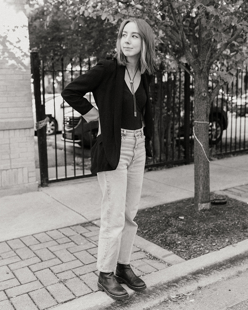 Amanda, wearing a blazer, high-waisted jeans, and a bolo tie, stands on a curb in Roger's Park with her hands positioned on her sides, looking to her right.